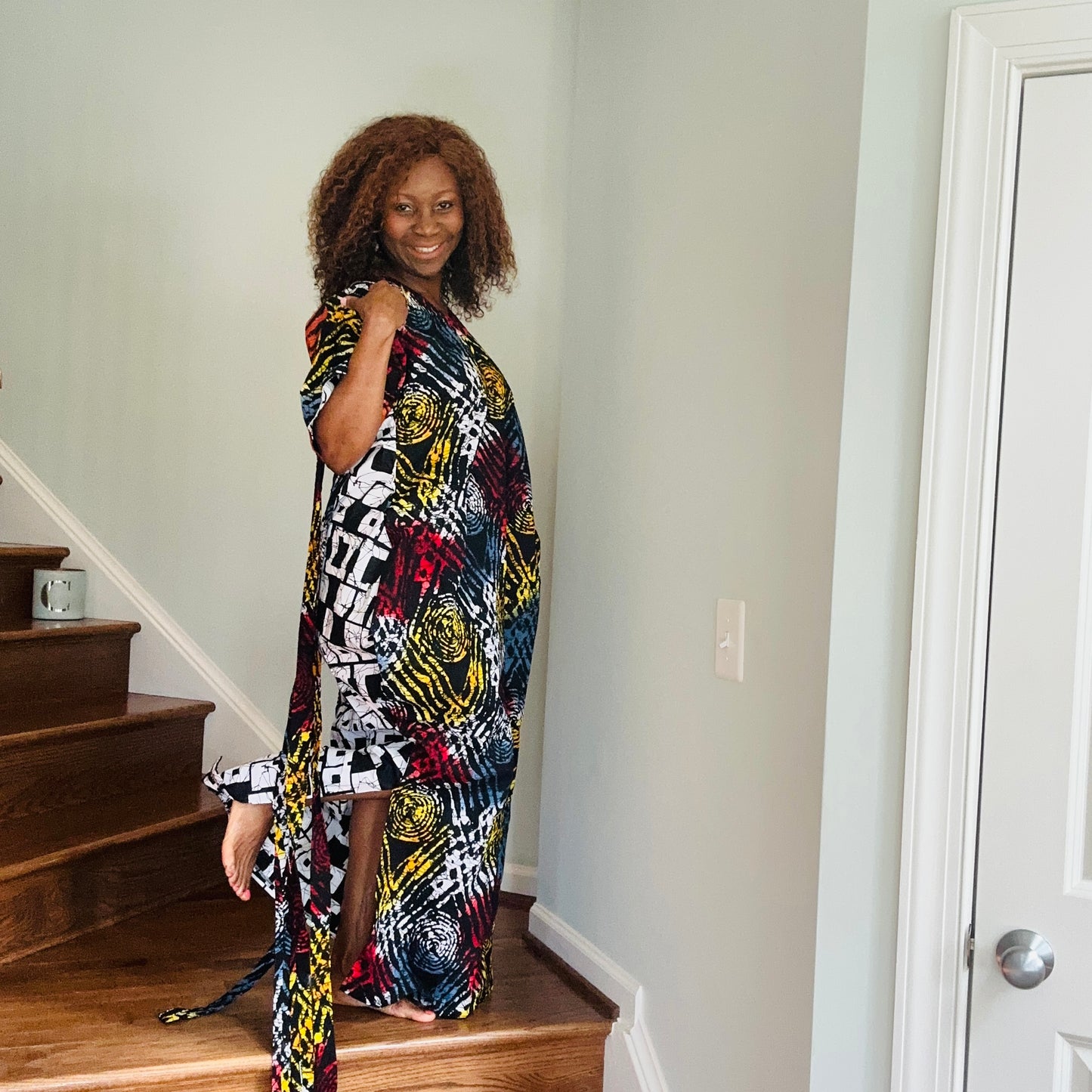 A woman standing on a staircase wearing a multicolored Ankara fringe dress with geometric patterns and tassel details.