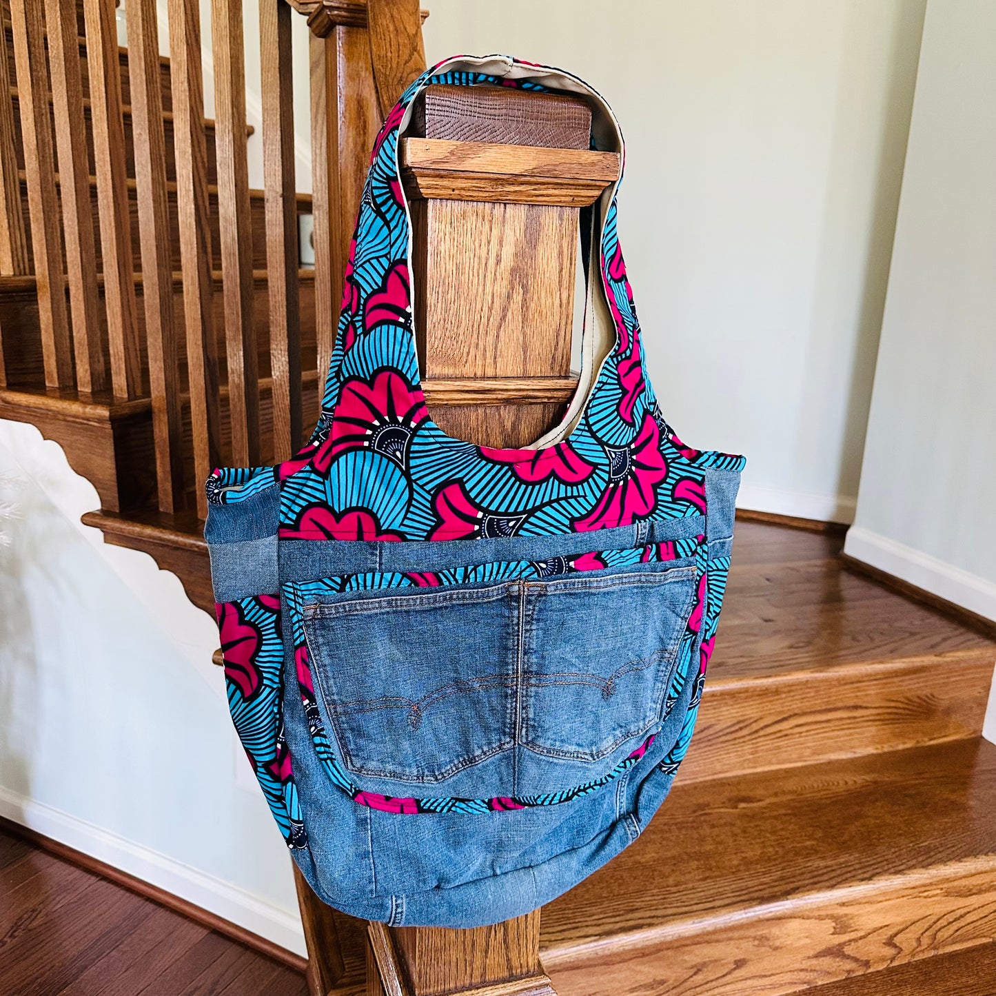 A denim bag with multicolored geometric patterns on the pockets and a denim pocket detail in front, displayed on a wooden hanger.
