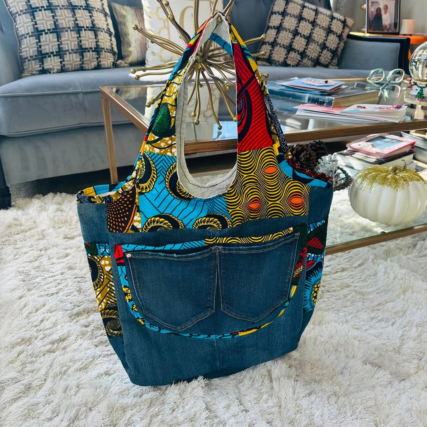 A denim bag with multicolored geometric patterns on the pockets and a denim pocket detail in front, displayed on a wooden hanger.